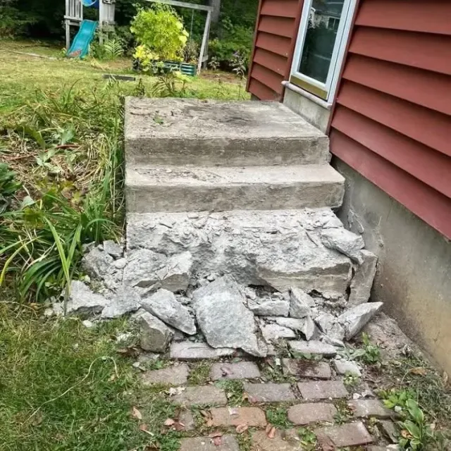 Crumbled concrete steps by a red house, with broken debris blocking the bottom stair.