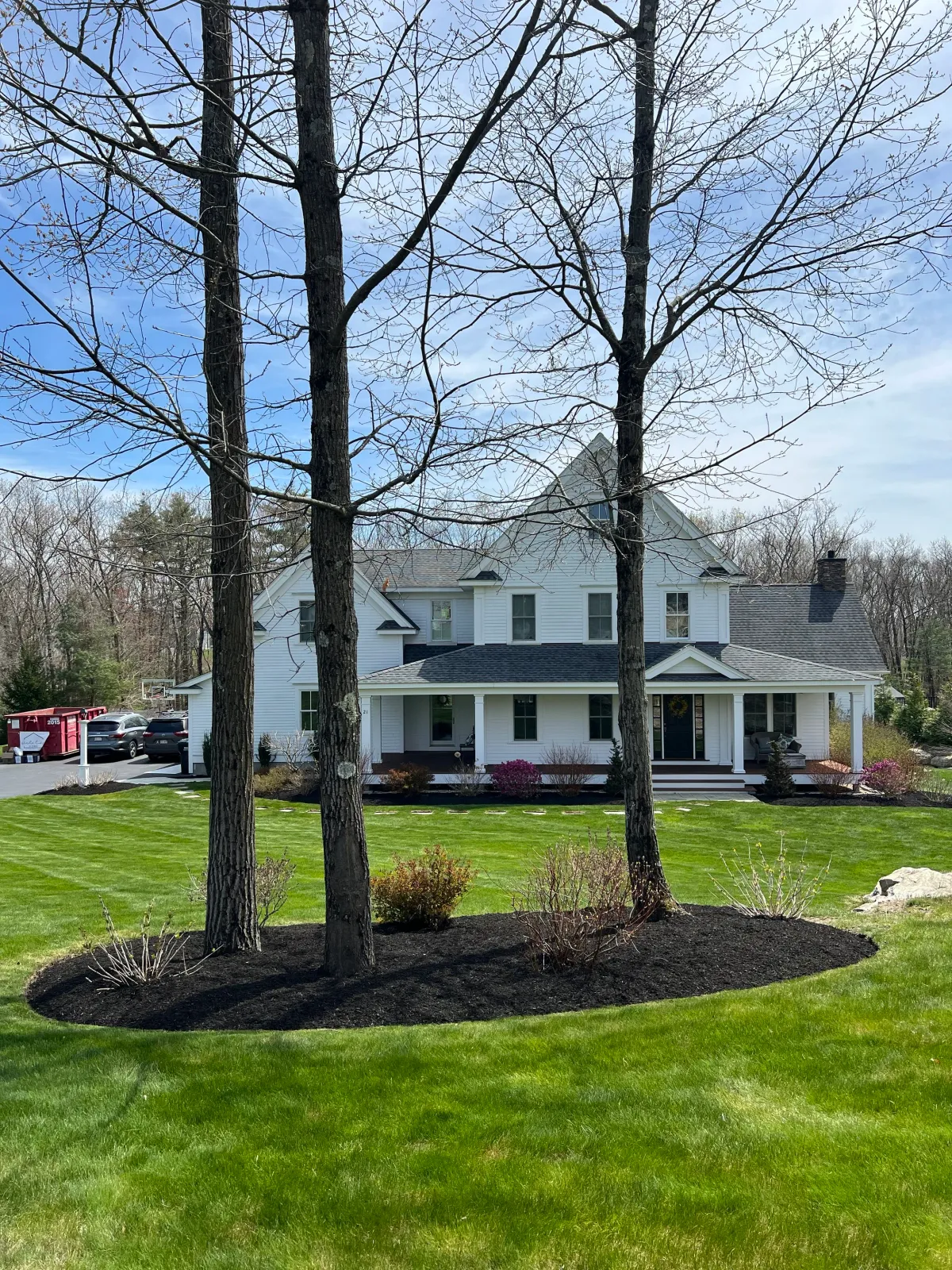 White two-story house with a dark roof, surrounded by a well-maintained green lawn and a landscaped flower bed with small shrubs and three large trees in the foreground.