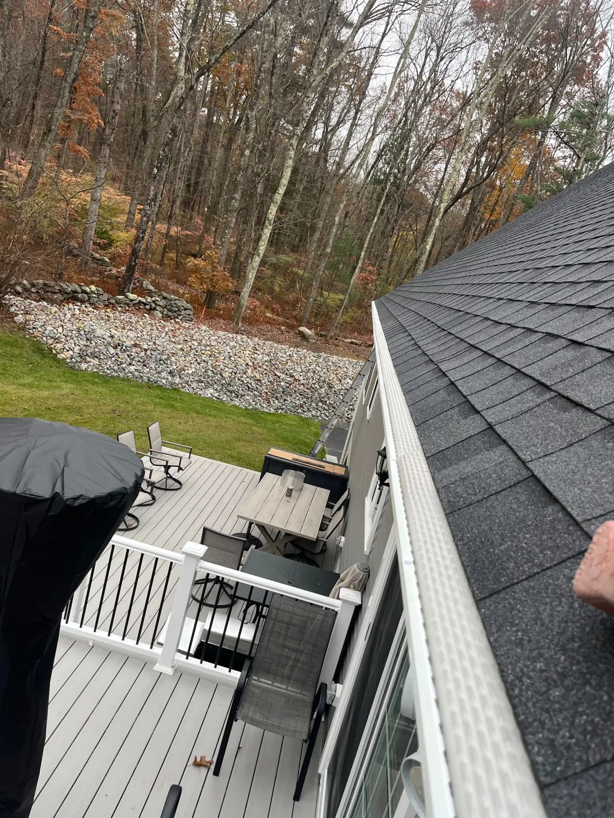 Backyard deck with white railing, black roof, patio furniture, and autumn trees beyond a rock border.