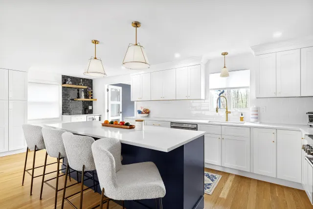 Modern white kitchen with navy blue island, white cabinets, hardwood floors, and gold fixtures. Three pendant lights hang above the island, which has four barstools. A window over the sink shows outdoor trees.