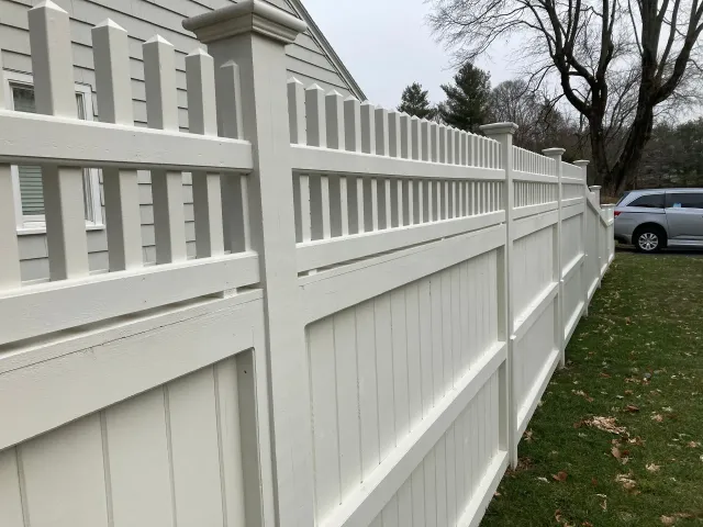 A white fence is surrounding a house with a car parked behind it.
