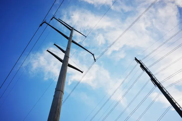 Two wooden utility poles with multiple crossarms and power lines against a blue sky with white.