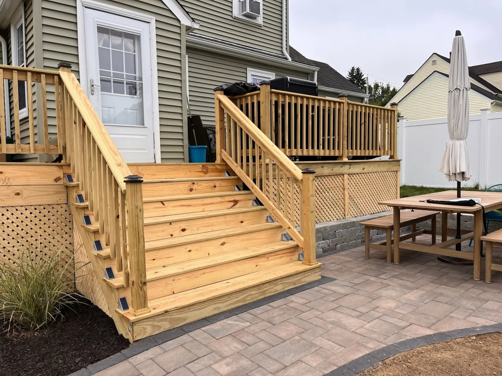 New wooden deck with stairs and railing attached to the back of a house, with a patio table, umbrella, and surrounding yard area.