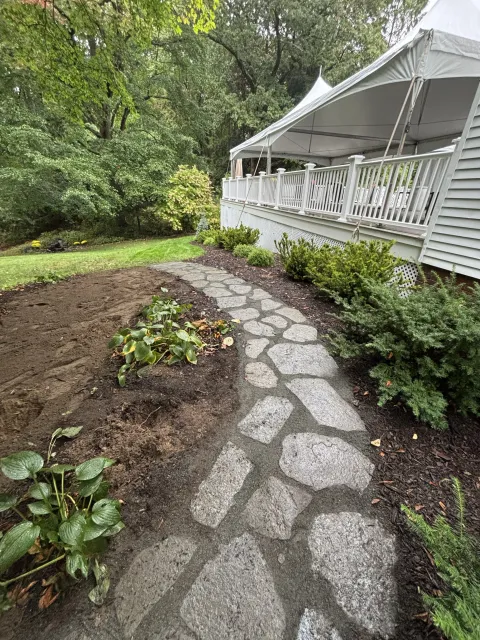 Stone pathway leading to a house with a porch, surrounded by greenery and a garden bed with plants.