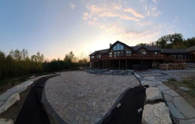 A large wooden house with a paved stone patio under a sunset sky, surrounded by trees and decorative boulders.