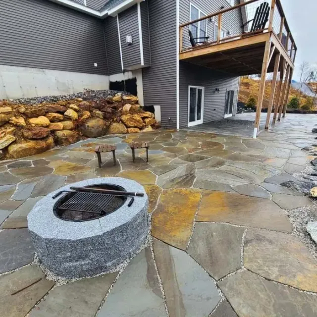 A gray stone fire pit sits on a flagstone patio beside a house with a wooden deck and a stacked rock retaining wall.