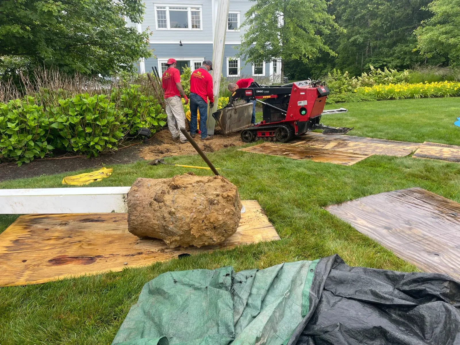 Workers in red shirts installing a large object in a residential garden using machinery, with plywood sheets protecting the grass.
