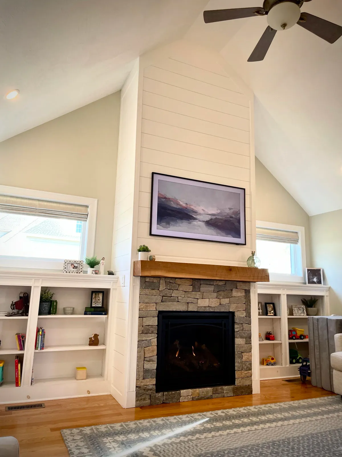 Living room with a stone fireplace, mounted landscape painting above, white built-in shelves with books and decor, windows on each side, hardwood flooring, patterned area rug, ceiling fan.