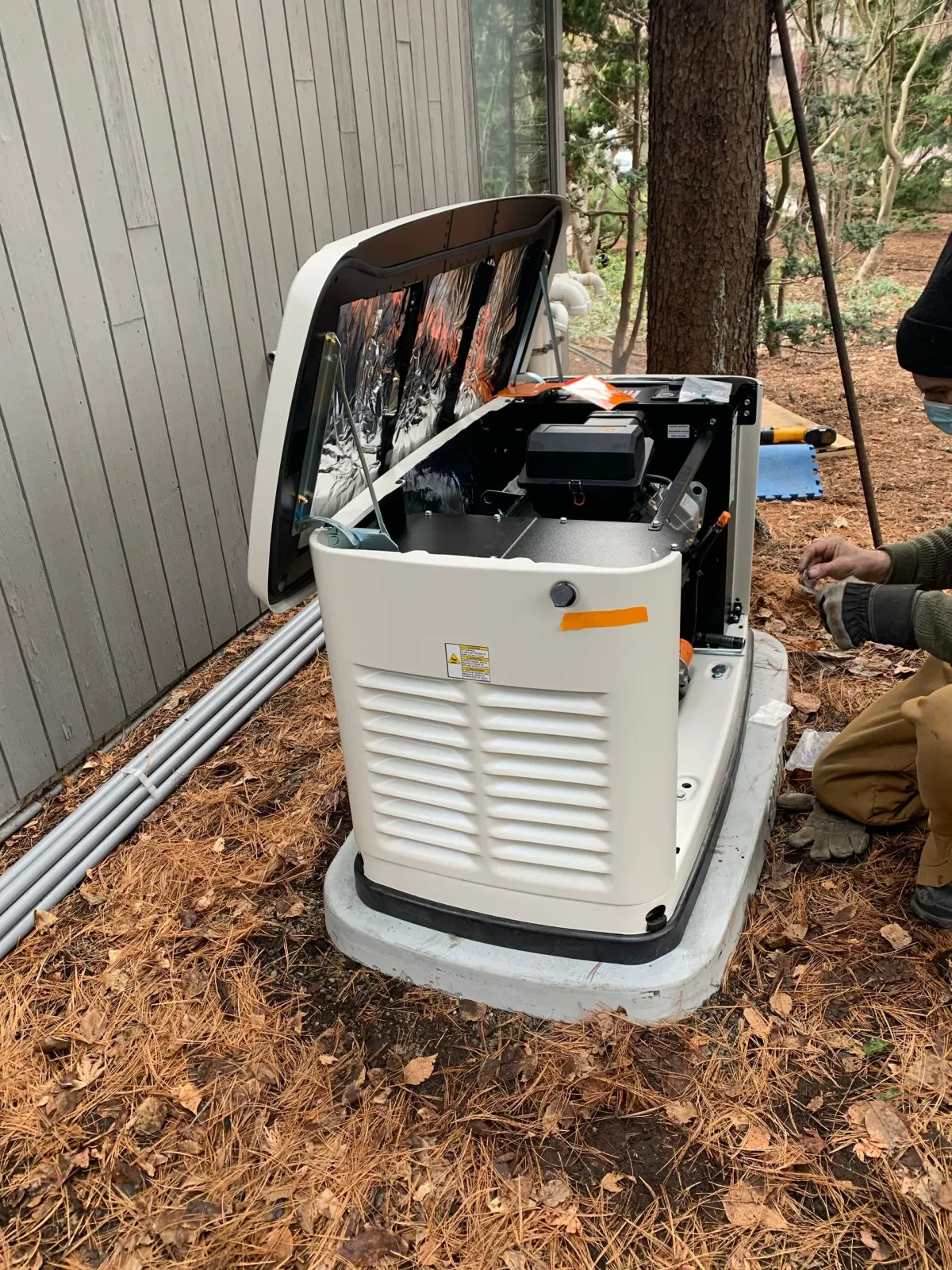 Generator with open access panel, being serviced by a person outdoors near a building and trees.