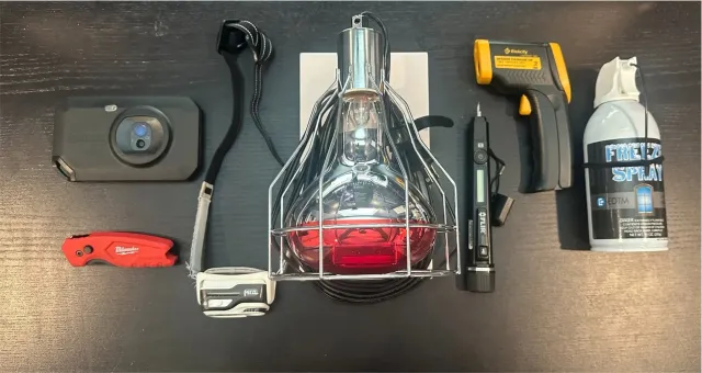 Collection of tools and equipment on a dark wooden surface, including a battery pack, a headlamp, a thermal scanner, a spray bottle labeled 'Freeze Spray', a utility knife, a lanyard, and a small electronic device with a screen.