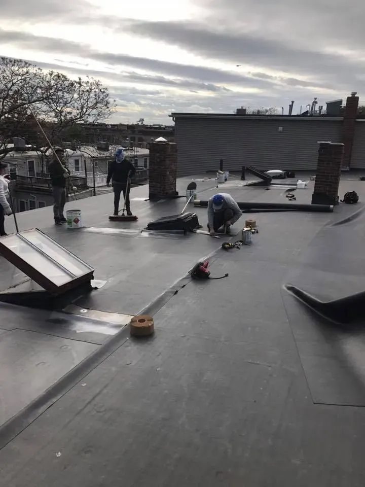 Workers installing a black flat roof on a building; some are bending over, others are standing. Overcast day.