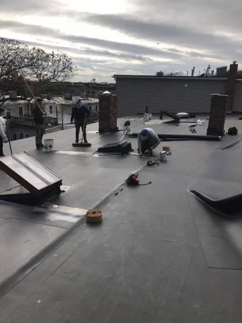 Workers installing a black flat roof on a building; some are bending over, others are standing. Overcast day.