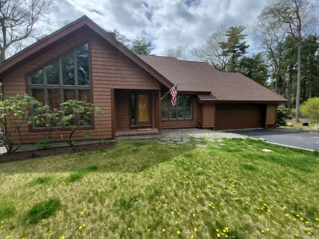 Brown house with large front window, garage, and American flag in a grassy yard under trees