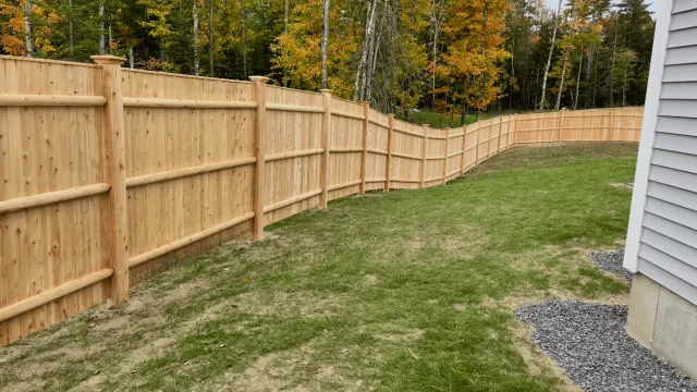 A newly installed wooden fence surrounds a grassy backyard with some patches of dirt, next to a house with white siding, with a wooded area in the background.