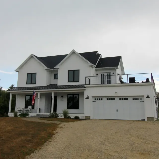 A modern white multi-story house with black roof and trim, front porch with chairs, and a gravel driveway, under an overcast sky.