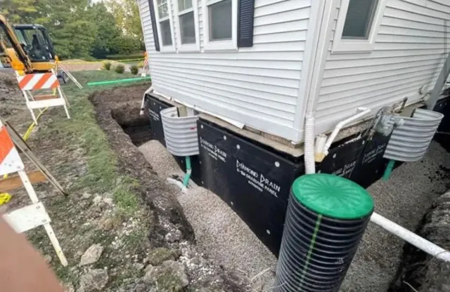 House foundation under excavation beside gray siding, with exposed black waterproofing and utility pipes