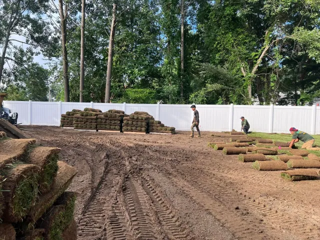 Three people work to lay rolls of sod in a muddy backyard in front of a white privacy fence and a stack of turf.