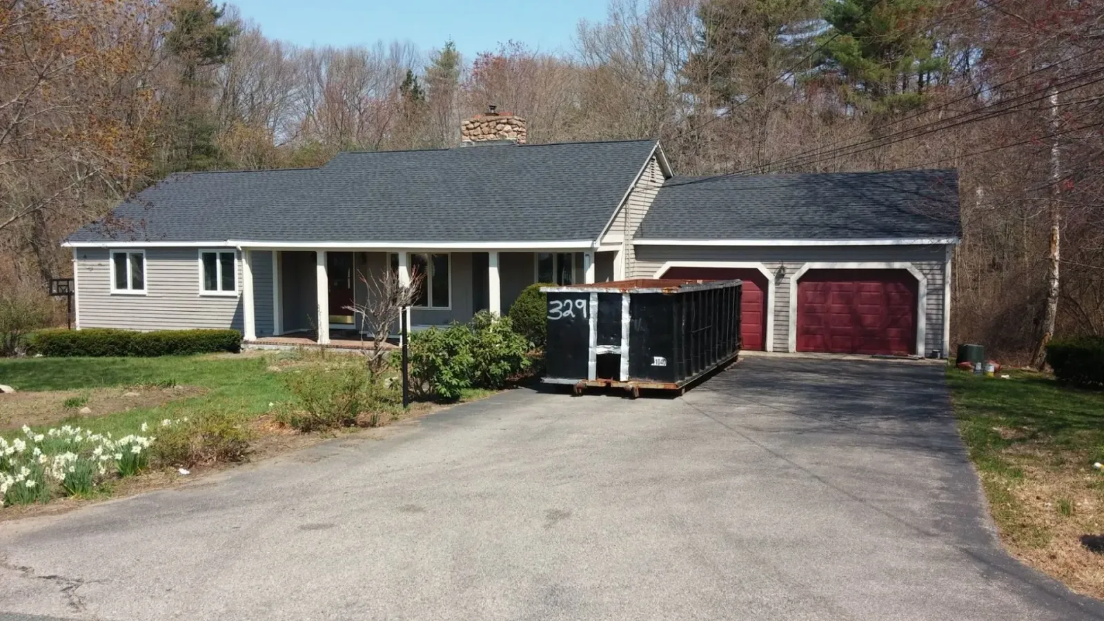 Ranch-style house with dark gray roof, two-car garage, and dumpster in driveway.