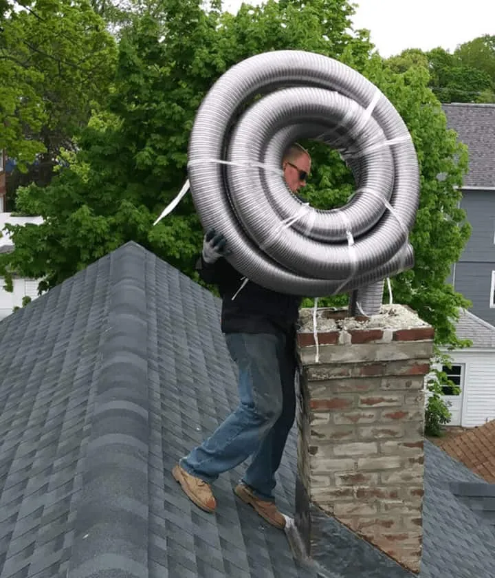 A man stands on a sloped roof in RI next to a brick chimney, carrying a large, coiled metal duct over his shoulders. Green trees and neighboring houses provide the background. Perfect for Chimney Repair Providence County needs.