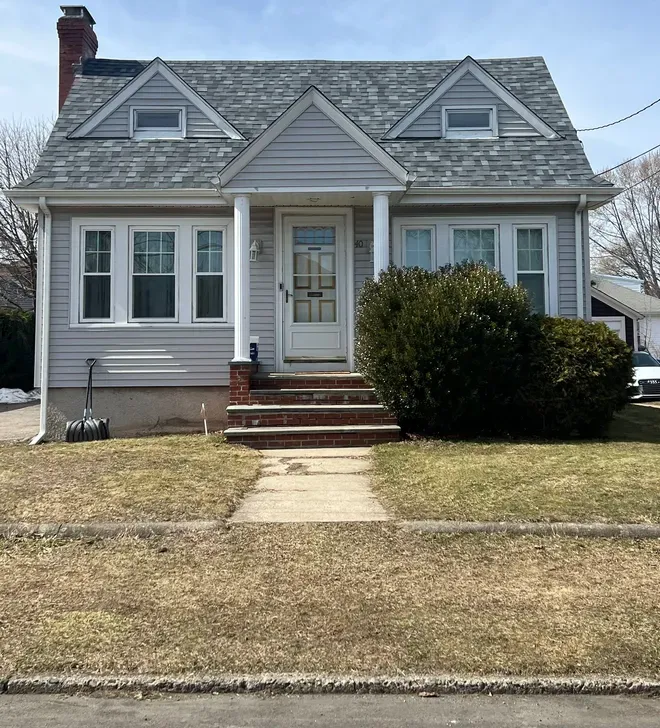 Small gray house with white trim, front porch, and brick steps behind a lawn and sidewalk.