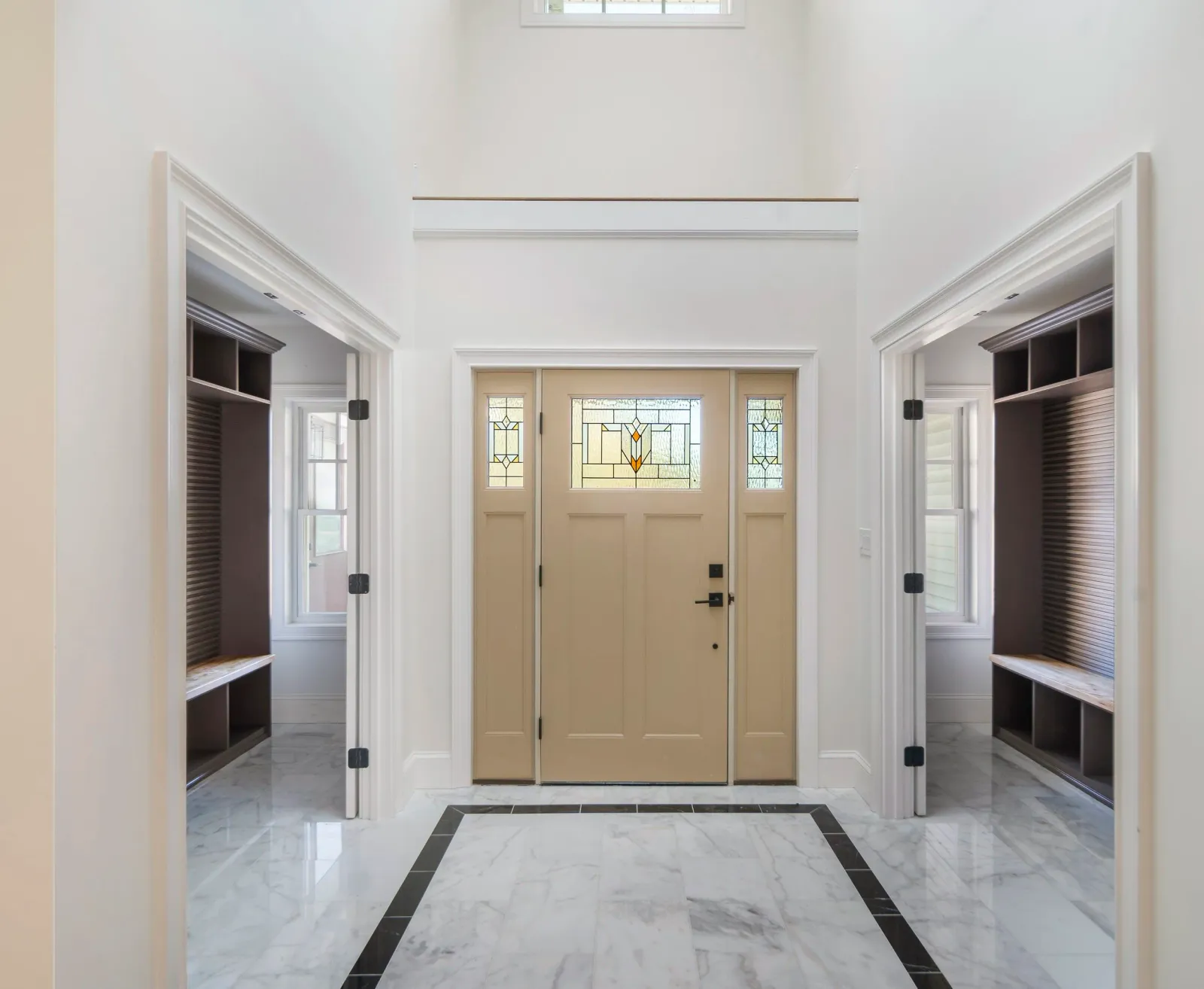 Entryway with a central beige door featuring stained glass panels, flanked by two side windows, and two open built-in cubby storage areas with dark wood finish, situated on marble flooring with a black border.