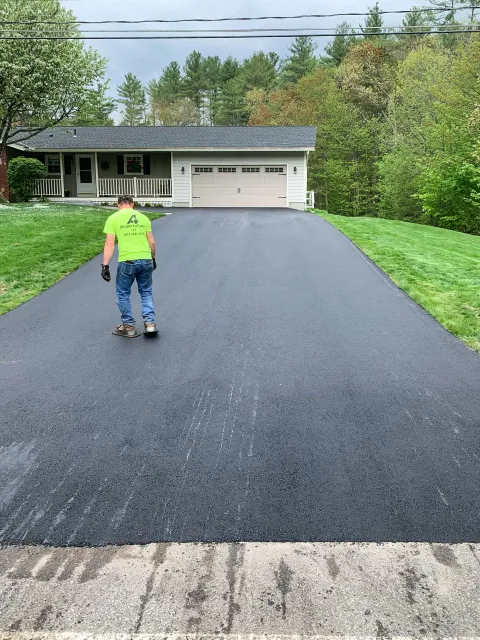 A worker in a neon green t-shirt and jeans roller-skating on a freshly paved asphalt driveway in front of a house with a garage, surrounded by green trees and lawn.