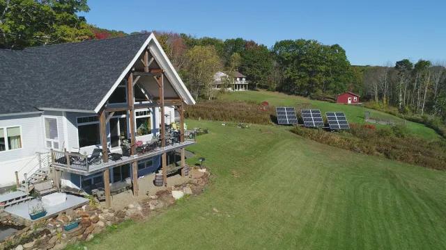 A white house with a dark gray gabled roof, large wooden supports, and a wraparound deck with outdoor furniture, surrounded by green grass and trees, with solar panels on a hillside and a small red shed in the distance under a clear blue sky.