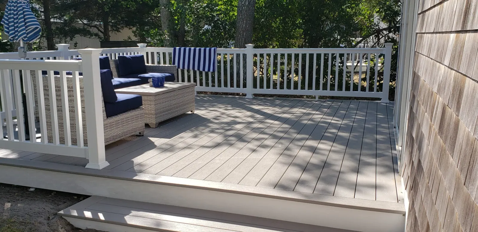Outdoor deck with wicker furniture and blue cushions, a striped towel hanging on the white railing, and a shaded area from surrounding trees.