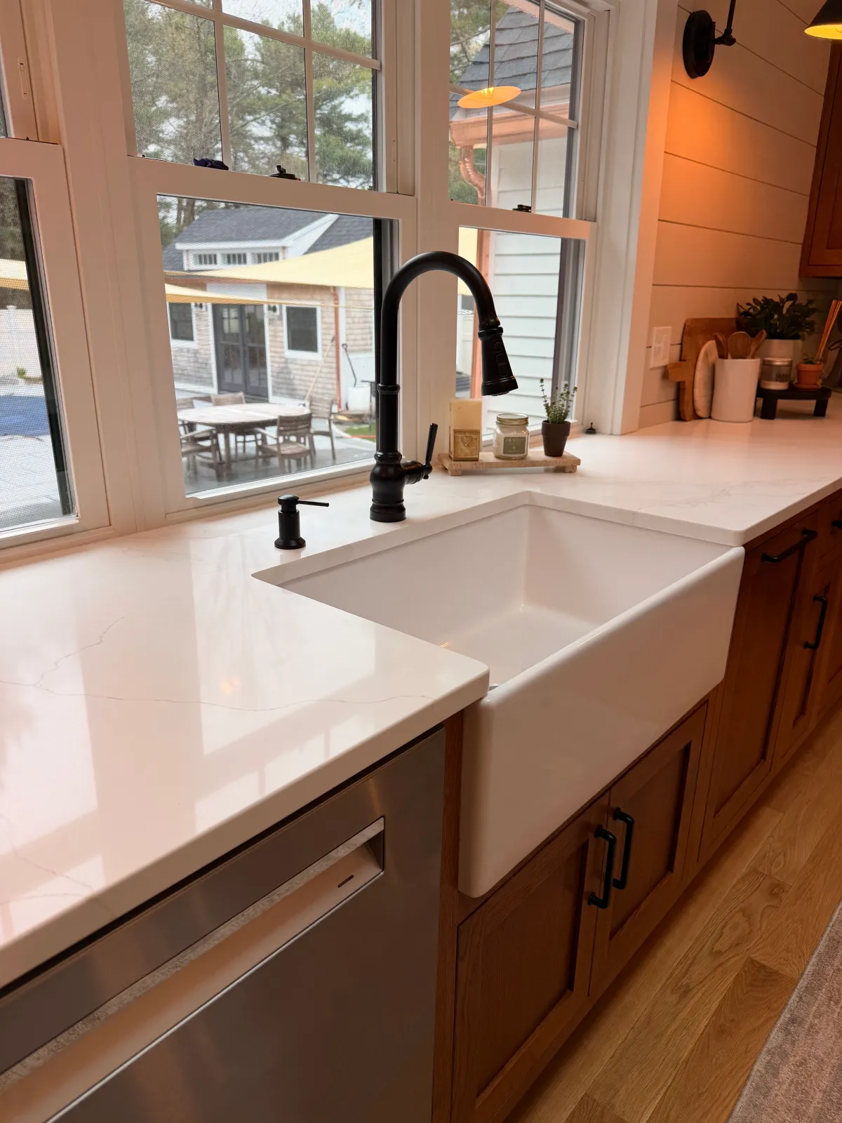Kitchen sink with black faucet, window above with outdoor patio furniture visible, copper-colored wood cabinets, beige countertop, small potted plant, candles on tray, wall-mounted speaker, and warm lighting.