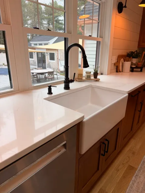 Kitchen sink with black faucet, window above with outdoor patio furniture visible, copper-colored wood cabinets, beige countertop, small potted plant, candles on tray, wall-mounted speaker, and warm lighting.