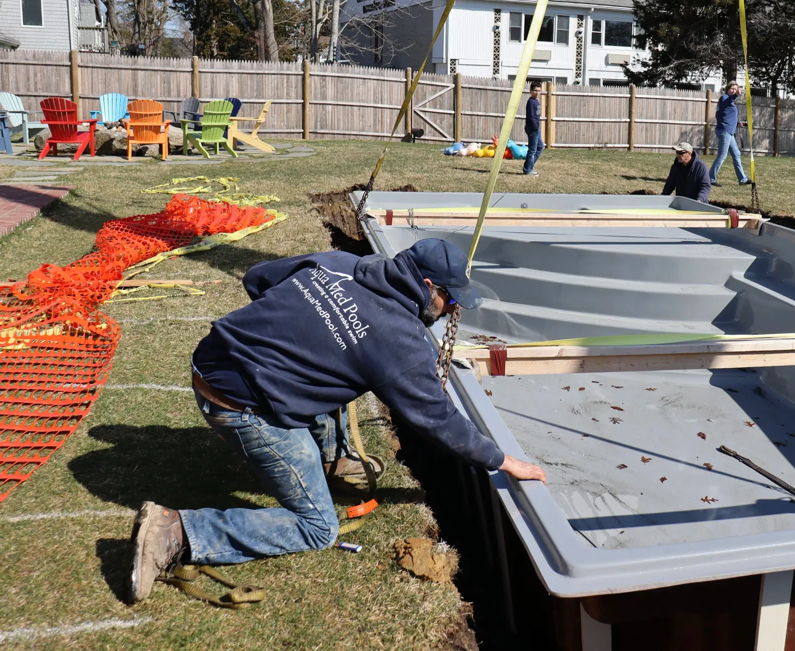 Workers installing a new inground swimming pool in a backyard, with orange safety fencing and colorful outdoor chairs nearby.