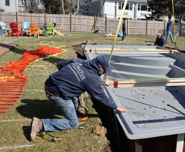 Workers installing a new inground swimming pool in a backyard, with orange safety fencing and colorful outdoor chairs nearby.