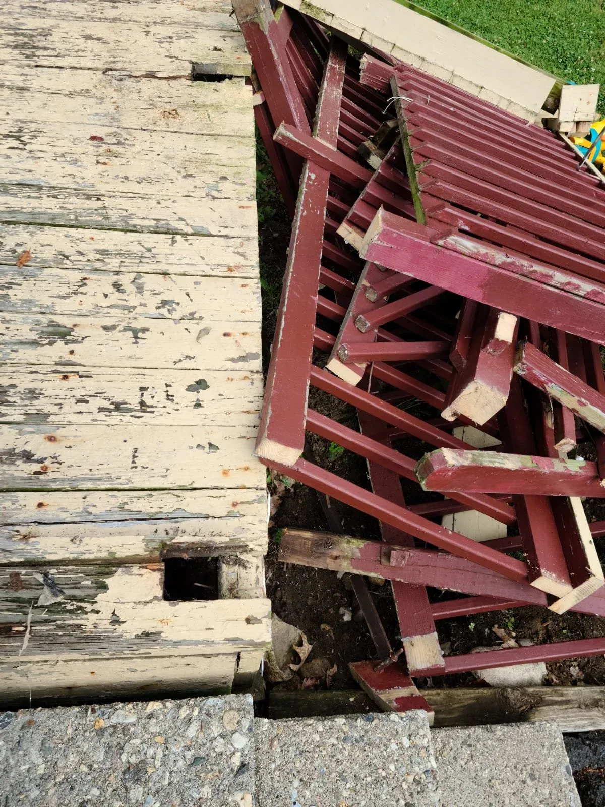 Close-up of a wooden porch with chipped paint next to a disassembled red-painted wooden structure, possibly a deck or railing, with some parts leaning against it.