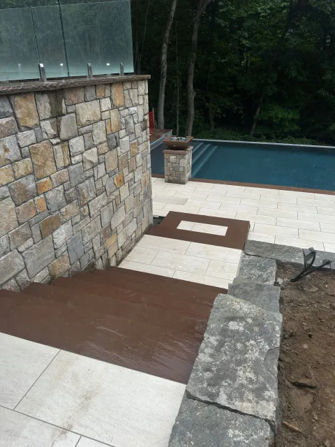 Stone patio with steps, a retaining wall, and a swimming pool beside a wooded area