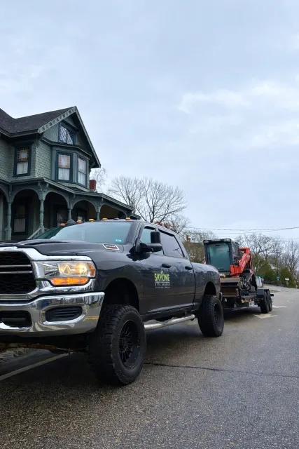 A black pickup truck with a logo and phone number on the door, towing construction equipment on a flatbed trailer, parked on a street in front of a Victorian-style house with blue-green paint and intricate woodwork