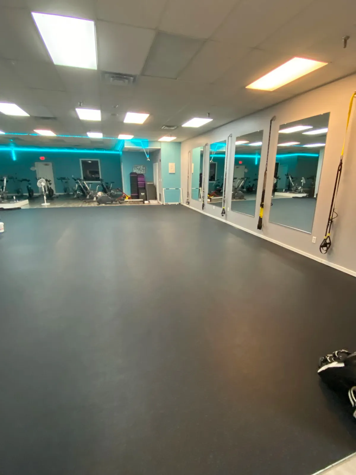 Empty fitness studio with black flooring, mirrors on the wall, and exercise equipment in the background, illuminated by ceiling lights and blue accent lighting.