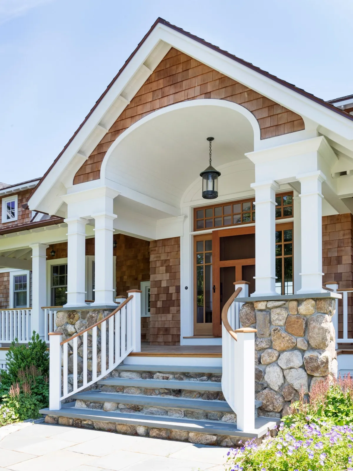 Front porch of a house with stone steps, white railings, wooden and shingle exterior, stone columns, and a large lantern light.