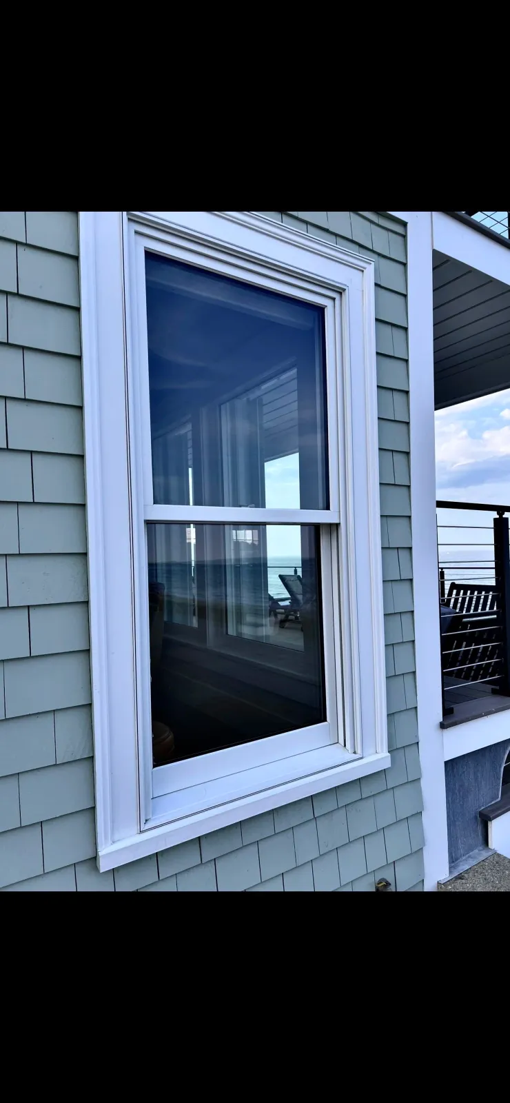 A window on a light green shingle house with a view of a balcony and the ocean in the background.
