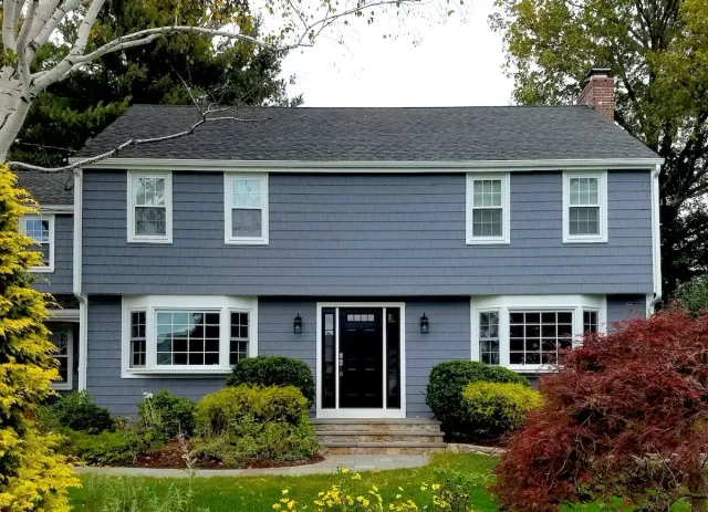 Two-story blue house with white trim, bay windows, black door, and landscaping.
