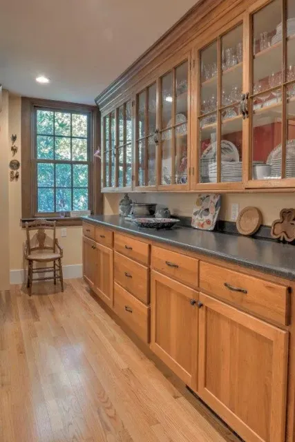 Long wooden kitchen with glass-front cabinets, dark countertops, and a window at the end.