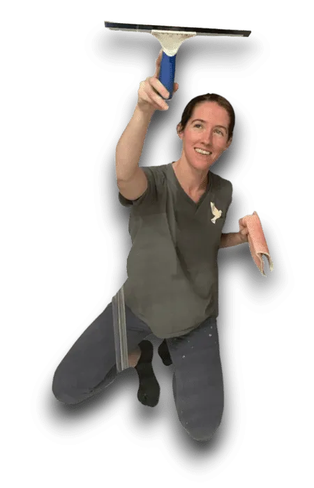 A woman kneels while cleaning a window with a squeegee and a pink cloth. She is smiling and wearing a gray t-shirt and blue pants, likely working on home cleaning tasks.
