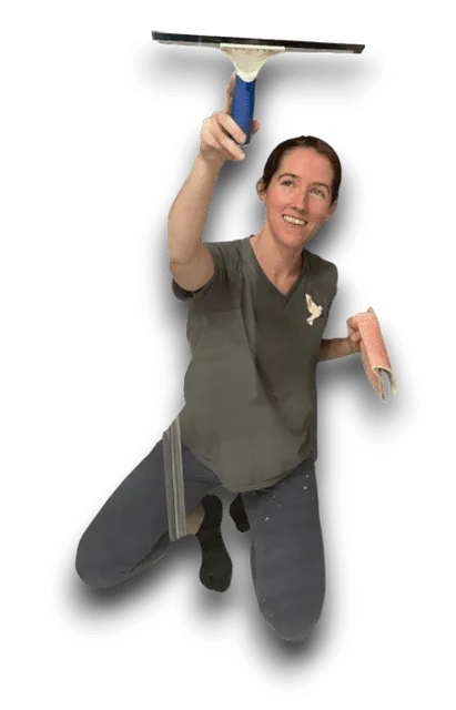 A woman kneels while cleaning a window with a squeegee and a pink cloth. She is smiling and wearing a gray t-shirt and blue pants, likely working on home cleaning tasks.