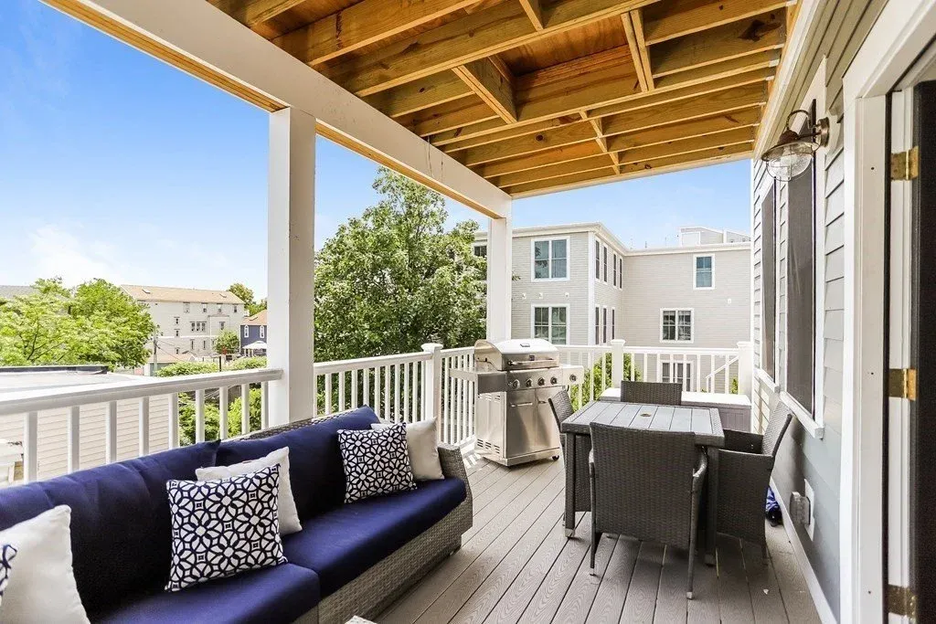 Covered porch with outdoor furniture, including a navy blue sofa with patterned throw pillows, a dining table with four chairs, a stainless steel grill, and a wooden ceiling, overlooking neighboring houses and trees.