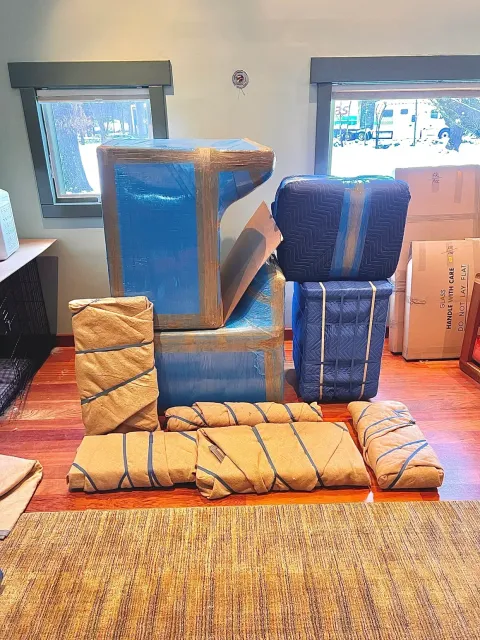 Packed furniture and boxes in a room with windows, wrapped for moving.