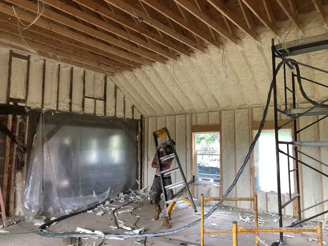 Installer in a respirator moving a stepladder along a vaulted ceiling line freshly sprayed with foam insulation