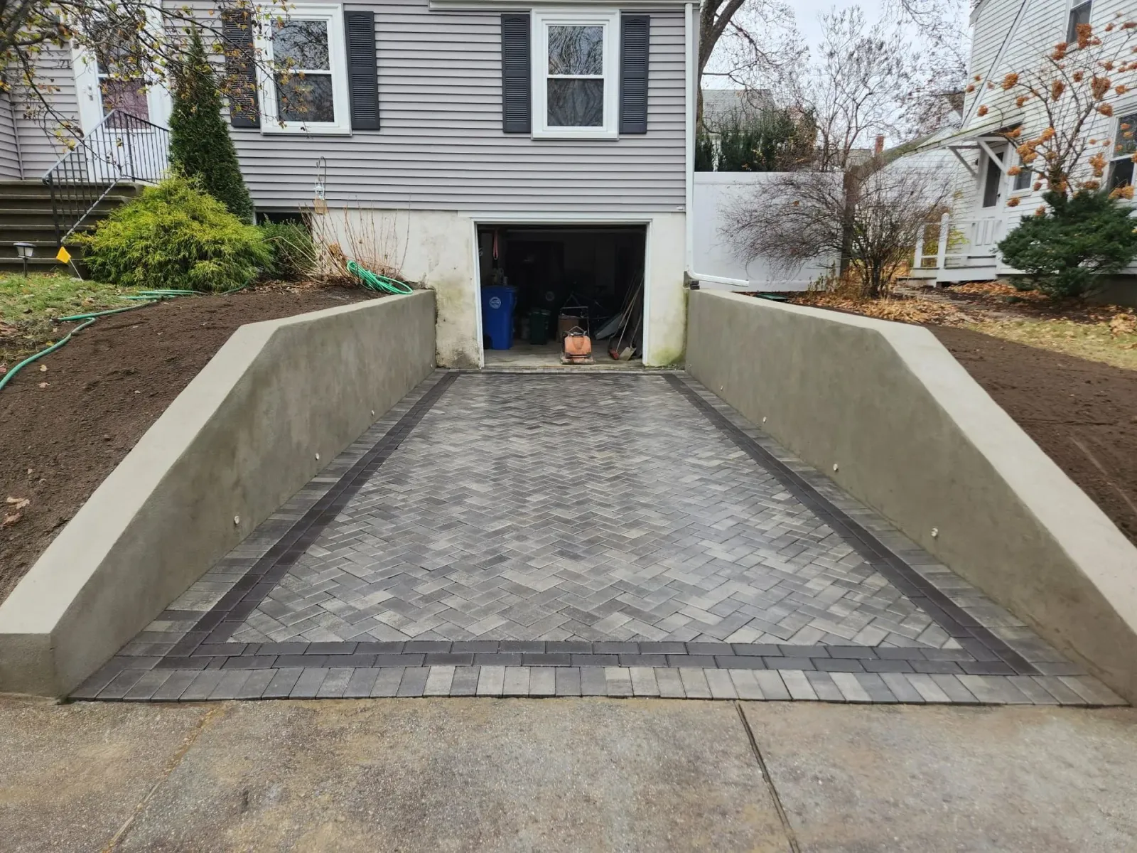 Concrete driveway descending to a garage under a house, bordered by low retaining walls.