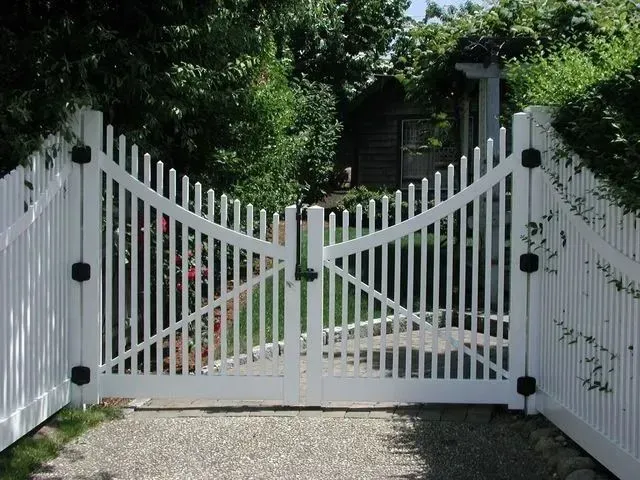 A white, double-swing wooden picket gate with a diagonal design at the entrance of a gravel driveway.