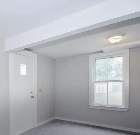 Small empty gray room with a white door, window, carpet, and ceiling light.