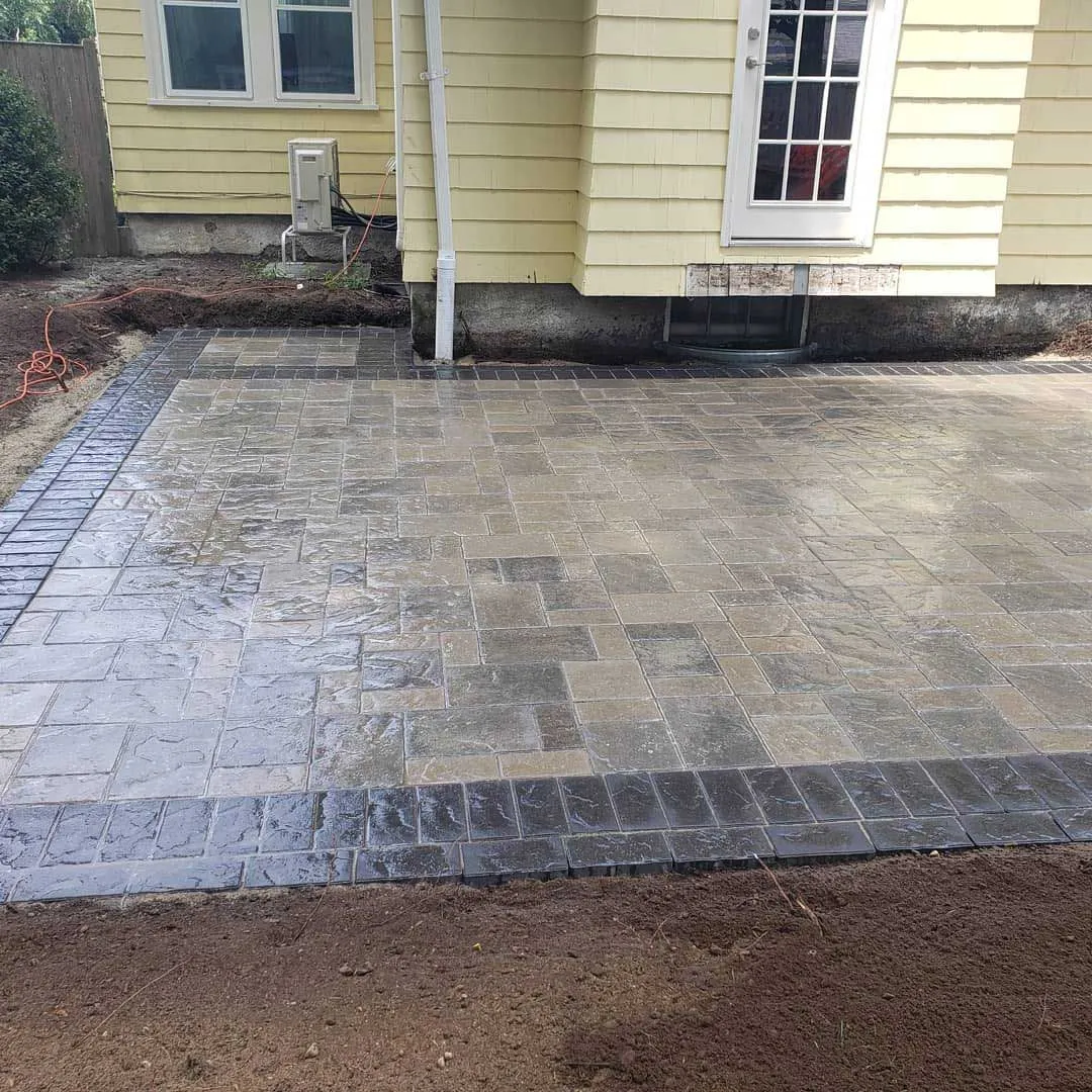 A stone patio with a light gray center and dark gray border sits beside a house with yellow siding and a white door.