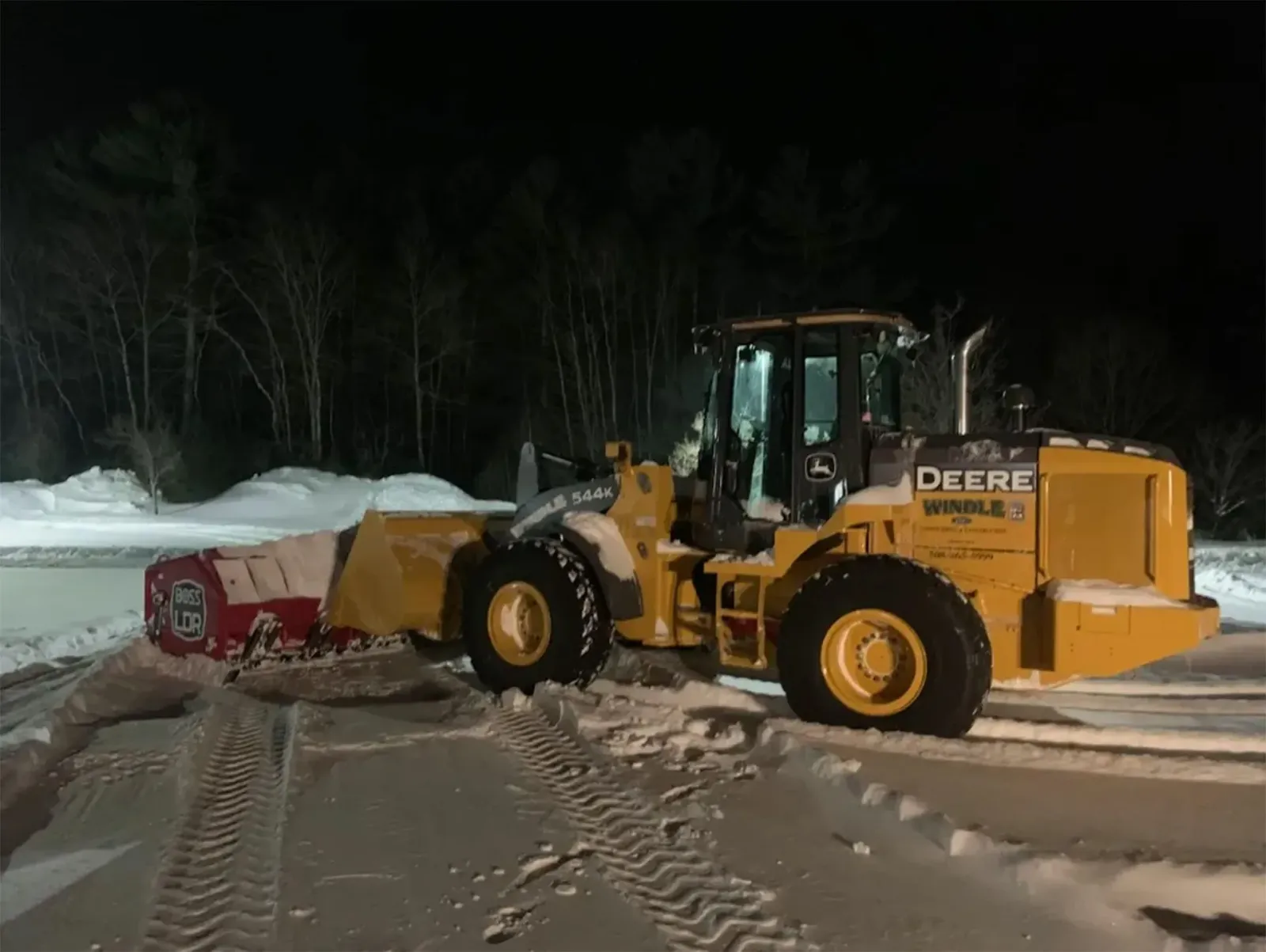 Yellow John Deere loader clearing snow beside a red truck at night